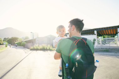 Sırt çantalı bir baba park kapısının önünde kucağında küçük bir kız tutuyor. Arka plan