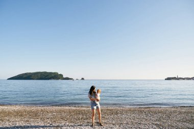 Annem bir çakıl taşı kumsalında duruyor ve kollarında deniz kenarında küçük bir kız tutuyor.