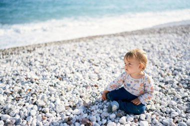 Çocuk deniz kıyısındaki çakıl taşlı bir plajda oturur ve uzaklığa bakar.