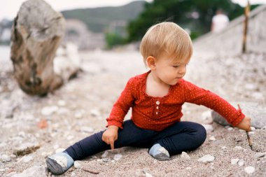 Çocuk çakıl taşı plajında oturur ve bir sopayla yere bir çukur kazar.