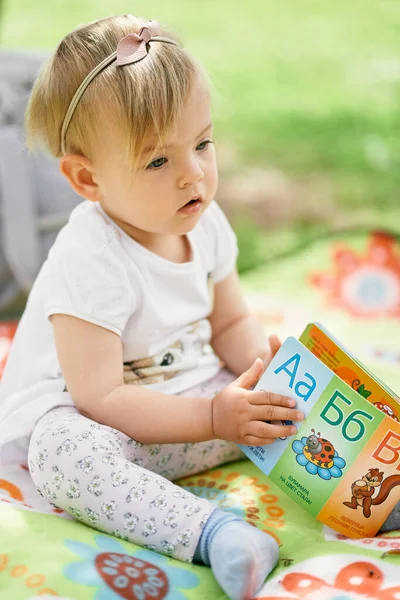 Gadis kecil duduk di atas selimut berwarna di halaman hijau dan memegang buku anak-anak di tangannya — Stok Foto