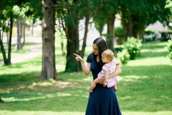 Anya egy kislányt tart a karjaiban, és a távolba vezeti a kezével. — Stock Fotó