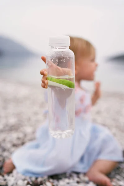Fles water met een schijfje citroen in de hand van een klein meisje in een jurk — Stockfoto