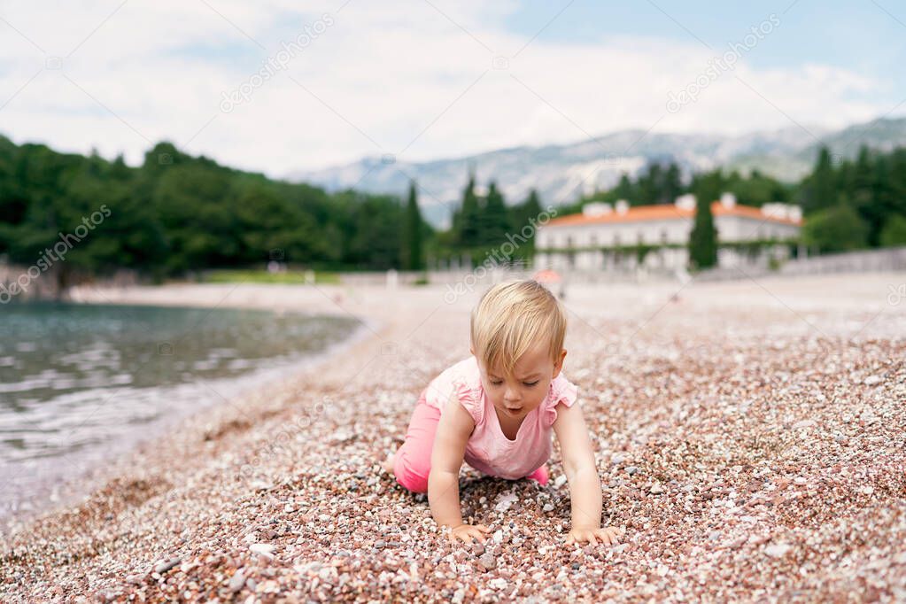 Little girl stands on her knees a pebble beach near the water. Villa Milocer