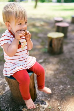 Küçük çıplak ayaklı bir çocuk bahçedeki bir kütüğe oturur ve bir kruvasan kemirir.