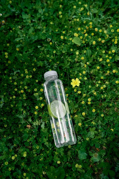 Бутылка воды с ломтиком лайма лежит на зеленой траве среди желтых полевых цветов — стоковое фото