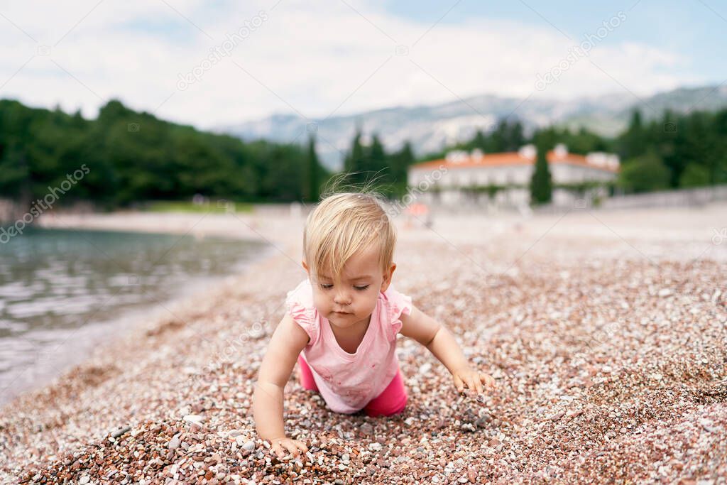 Little girl kneels on a pebble beach near the water