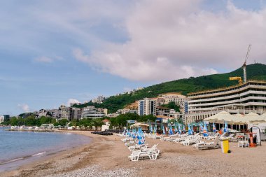 Katlanmış şemsiyeli güneş şezlongları, binaların, yeşilliklerin ve dağların zeminine karşı sahilde dururlar.