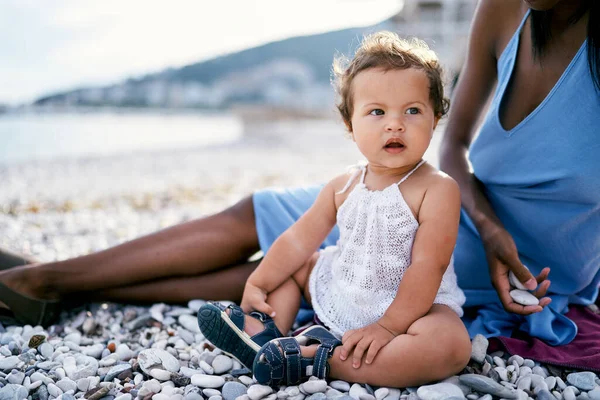 입을 벌린 어린 소녀 가손에 조약돌을 들고 어머니 옆 해변에 앉아 있다 — 스톡 사진