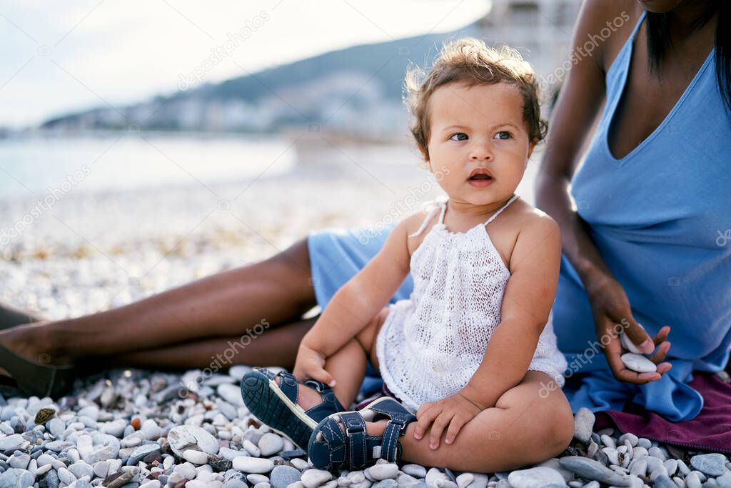 Little girl with an open mouth sits on the beach near her mother holding pebbles in her hand