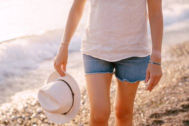 Şortlu bir kadın elinde şapkasıyla su kenarında bir çakıl taşı plajı boyunca yürüyor.