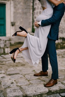 Damat, evin avlusundaki kaldırım taşlarının üzerinde dururken gelini kollarında tutuyor.