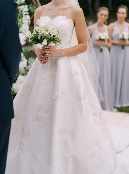 Sposa in un abito bianco con un mazzo di fiori tra le mani si trova accanto allo sposo sul prato — Foto Stock