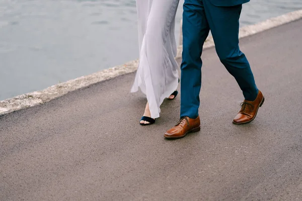 Legs of the groom in blue trousers and the bride in a white dress are walking along the asphalt road over the sea. Close-up — Stock Photo, Image
