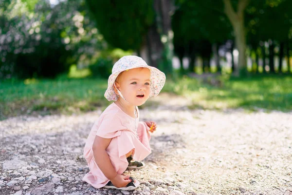 Liten flicka i en hatt huk på en grusgång i parken — Stockfoto