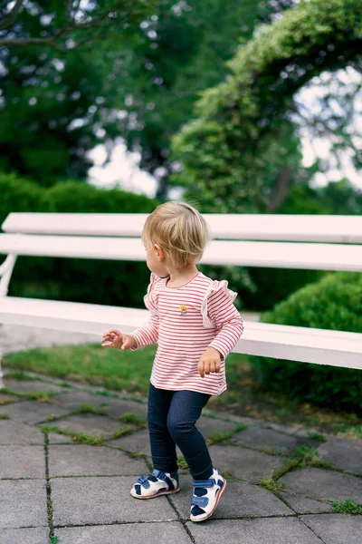 Little girl stands at a wooden bench in the park, turning her head back — Stock Photo, Image