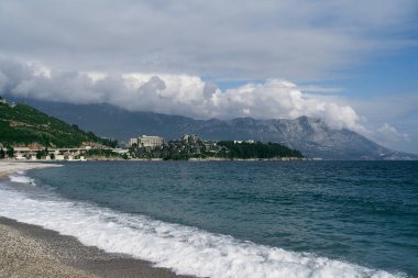 Deniz dalgaları çakıl taşlı sahilde dağların ve binaların arka planına doğru akar.