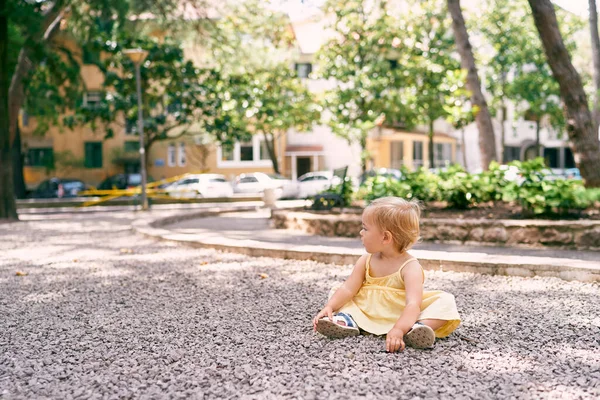 Liten flicka i klänning sitter på en grusgång på gården i huset — Stockfoto