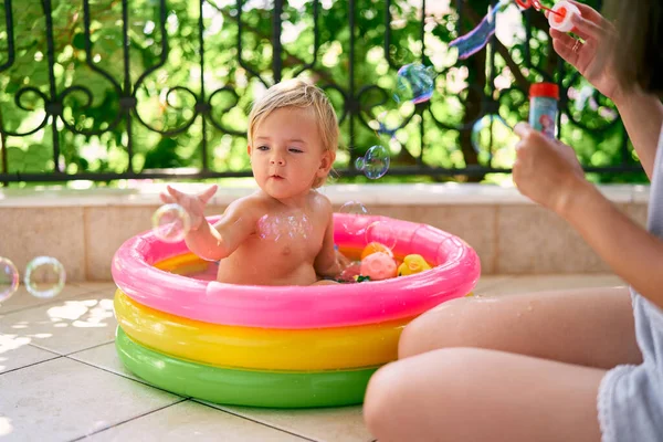 Mamma soffia bolle di sapone, e una bambina li cattura mentre si siede in una piscina gonfiabile — Foto Stock