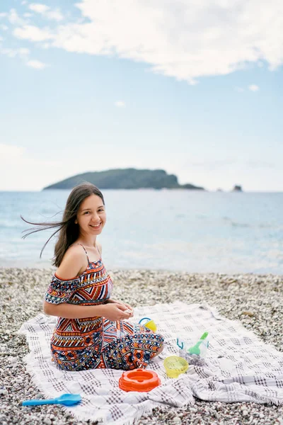 Smiling girl sits on a bedspread on a pebble beach with childrens toys — 스톡 사진