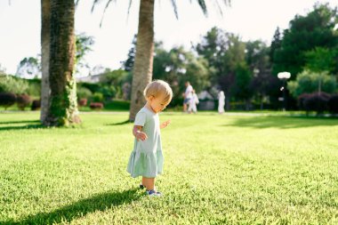 Küçük kız yeşil çimlerin üzerinde duruyor ve ayaklarına bakıyor. Yan görünüm