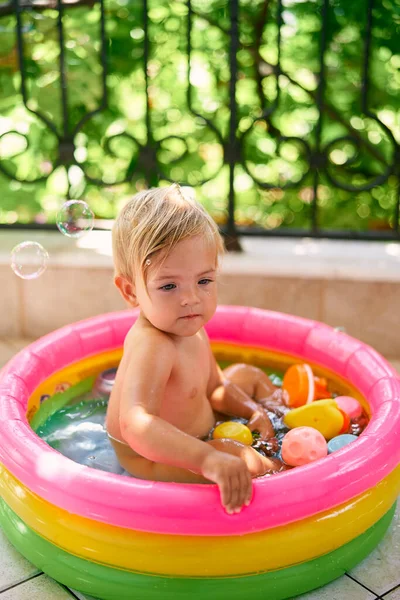 O bebê pequeno doce senta-se em uma piscina inflável pequena com brinquedos — Fotografia de Stock