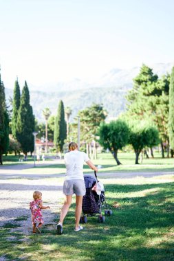 Yeşil parkta, yanında bebek arabasıyla dolaşan küçük bir kız.