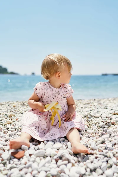 Little girl with a banana in her hand sits on a pebble beach, turning her head to the side — 스톡 사진