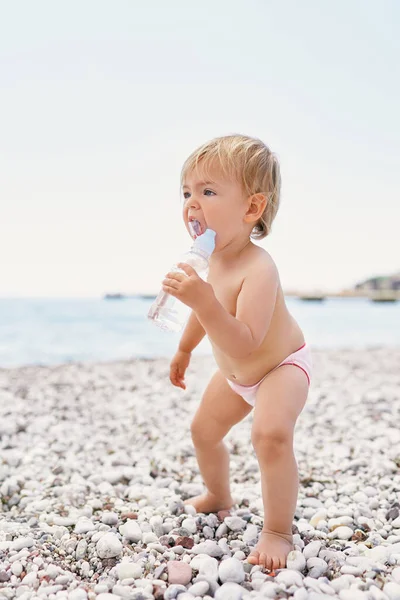 Small child in shorts stands on a pebble beach with a bottle of water in his hands — 스톡 사진