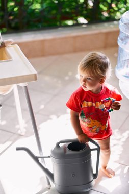 Elinde çıngıraklı bir çocuk su bidonunun yanındaki balkonda duruyor.