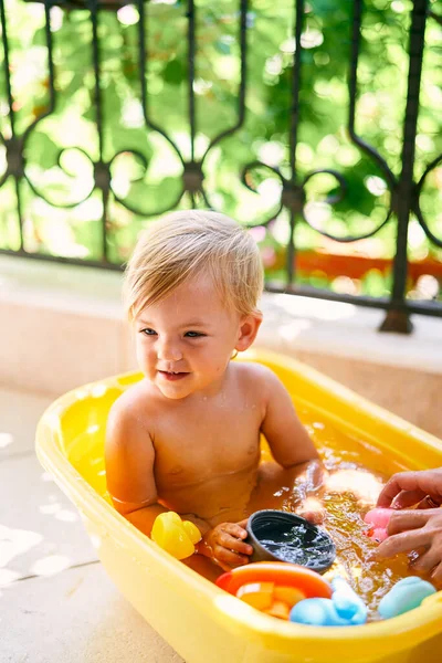 Bambino piccolo con una paletta tra le mani si siede in un bacino con acqua e giocattoli sul balcone — Foto Stock