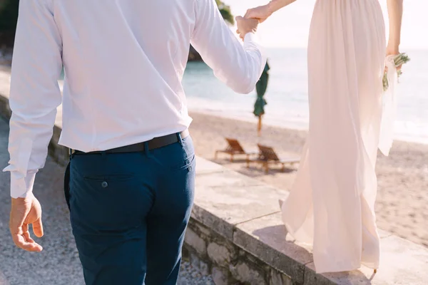 Ο γαμπρός κρατά το χέρι της νύφης με ένα μπουκέτο, περπατώντας κατά μήκος της παραλίας — Φωτογραφία Αρχείου
