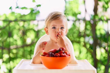 Kirli suratlı çocuk masada oturup kiraz yiyor.