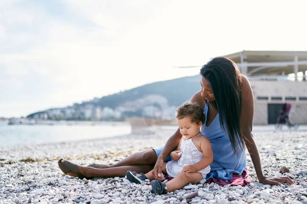 Ibu dengan seorang gadis kecil duduk di pantai dan menunjukkan kerikil padanya — Stok Foto