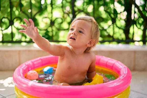 Carino bambino si siede in una piccola piscina gonfiabile con giocattoli e tira su la mano — Foto Stock