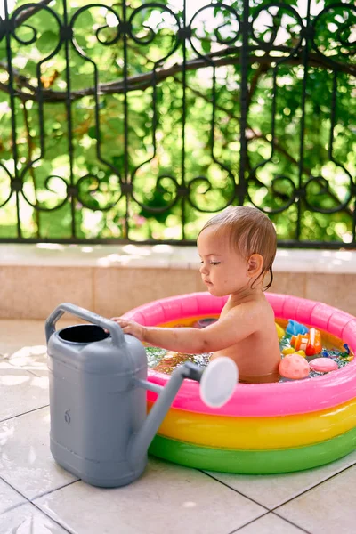 Il bambino si siede in una mini-piscina gonfiabile, accanto a un annaffiatoio da giardino — Foto Stock