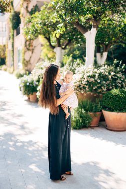 Annem küçük bir kızla kollarında yeşil çalılarla küvetlerin yanında duruyor.