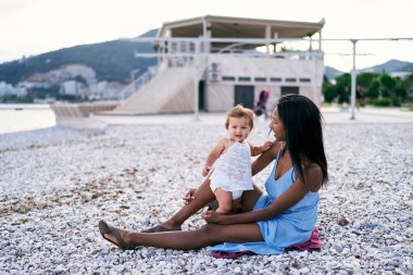 Annem deniz kenarındaki çakıl taşlı sahilde oturuyor. Yanında da küçük bir kız var.