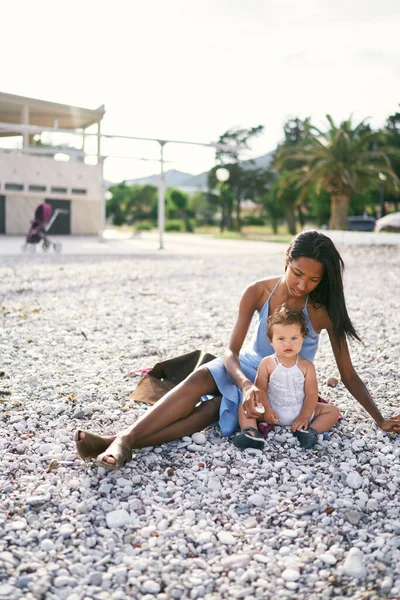 Ibu duduk di pantai dan memegang kerikil untuk seorang gadis kecil — Stok Foto