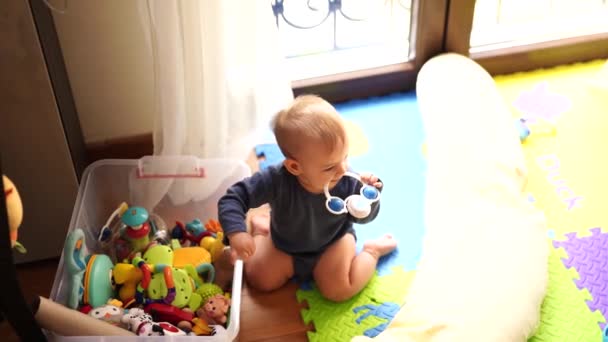 Kid senta-se no chão em um tapete colorido perto de uma caixa com brinquedos e rói um chocalho — Vídeo de Stock