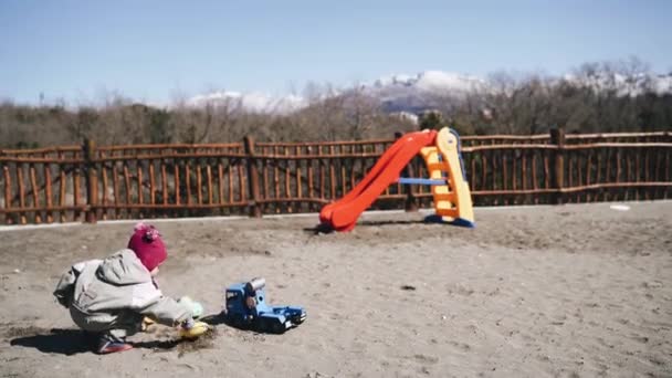 Gadis kecil menuangkan pasir dengan sekop ke dalam mobil mainan dekat slide — Stok Video
