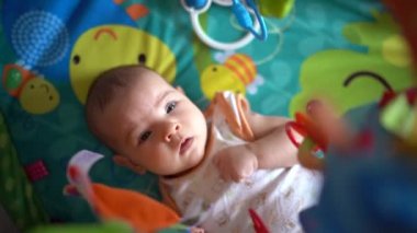 Yeni doğmuş bir bebek oyun hasırının üzerinde yatar ve asılı oyuncaklara bakar.