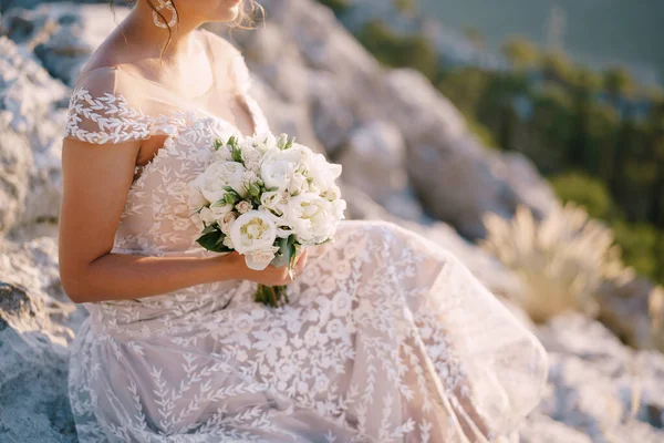 Bruid met een boeket pioenen zit op de rotsen tegen de achtergrond van bergen en groen — Stockfoto