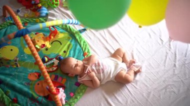 Yeni doğmuş bebek kollarına ve bacaklarına balonlar bağlanmış bir şekilde bir paspasın üzerinde yatıyor.