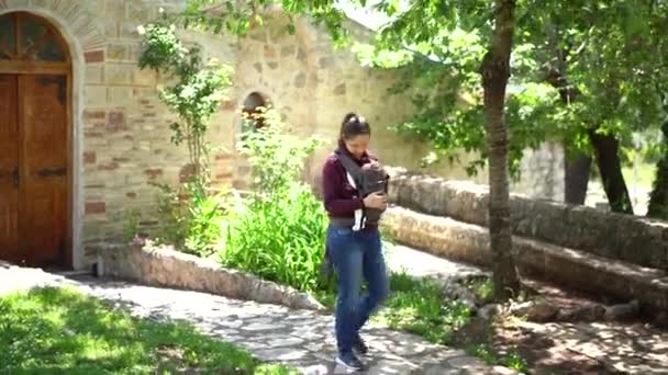 Mamma smeker en nyfödd i en lyftsele medan hon går längs stigen i parken — Stockvideo