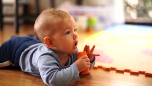 Kind bijt een wortel liggend op zijn buik op de houten vloer — Stockvideo