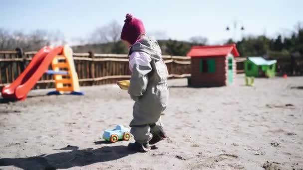 Menina derrama areia com uma espátula de brinquedo em um carro de brinquedo no playground — Vídeo de Stock