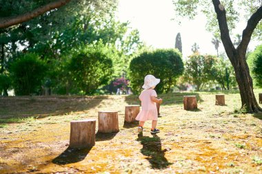 Şapkalı küçük bir kız çimler boyunca yürür, kütüklerin yanından geçer. Arka plan