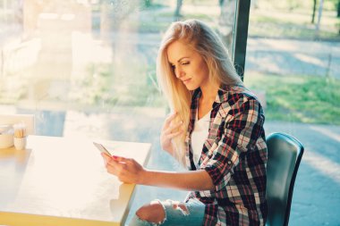 Genç kız öğrencinin cep telefonu ile modern kahve dükkanında oturmuş. Kafede pencerenin önünde oturan Sunset.Woman.