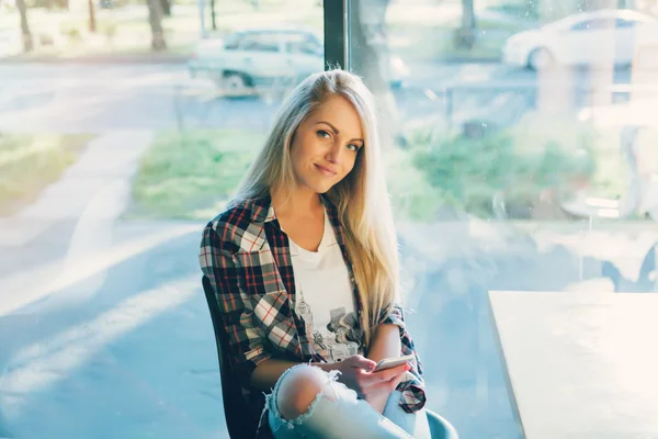 Jovem estudante sentada no café moderno com telefone celular. Pôr do sol. Mulher sentada na frente da janela no café . — Fotografia de Stock